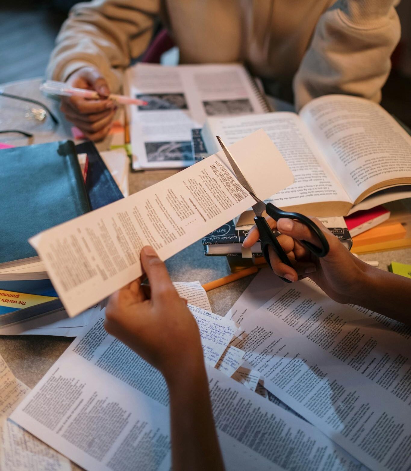 Two students involved in a study session with books, cutting paper, and taking notes.